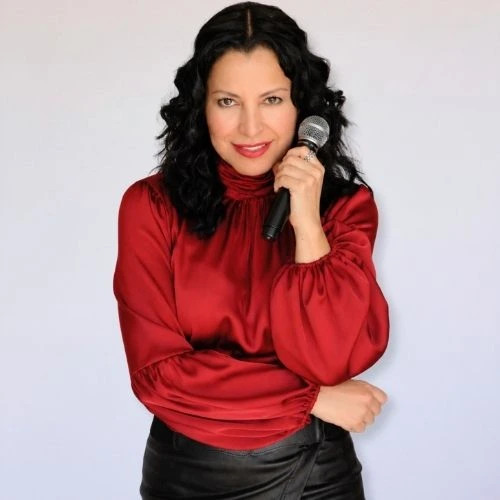 Stand-Up Comedian in Boca Raton Mariza Brussolo wearing a red blouse holding a microphone looking at the camera and smiling